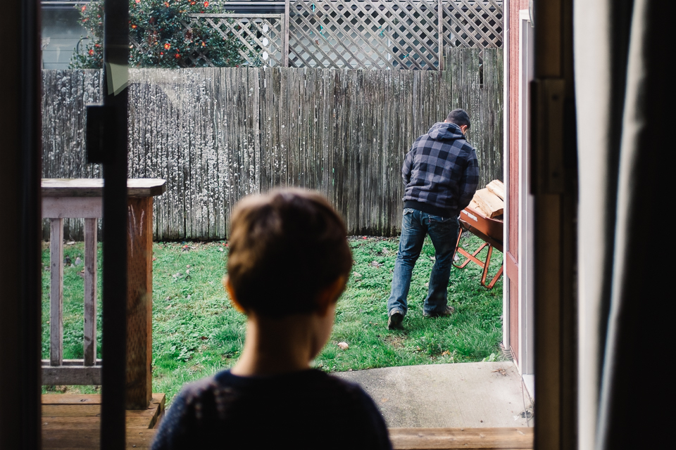 child watches father with wheel barrow through window -Documentary Family Photography