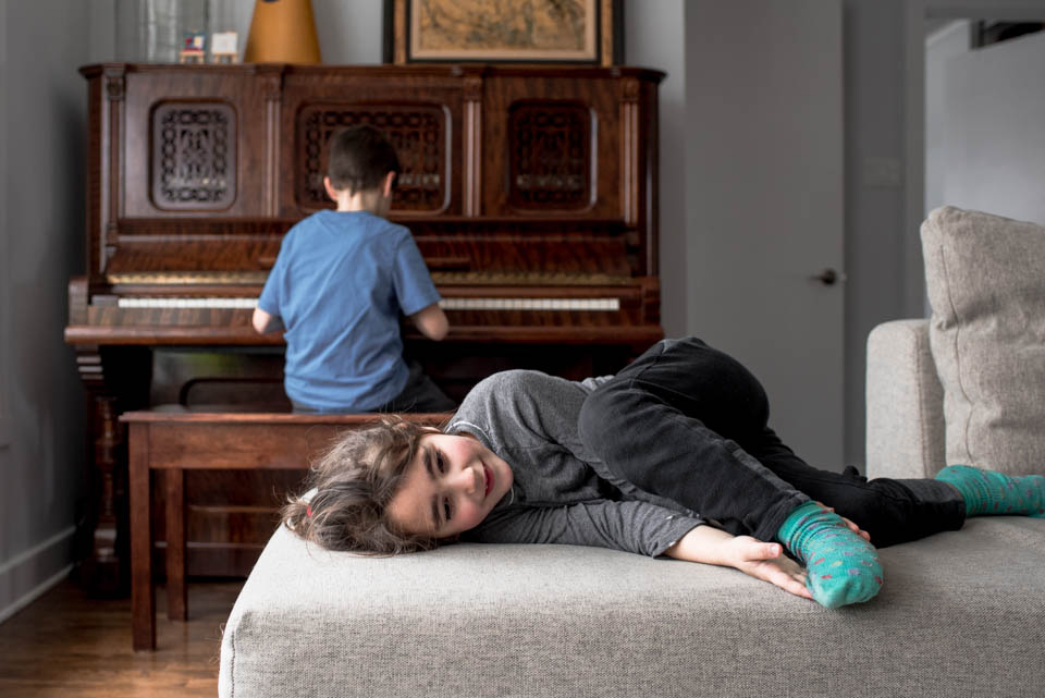 girl laying on ottoman while boy plays piano - Documentary Family Photography