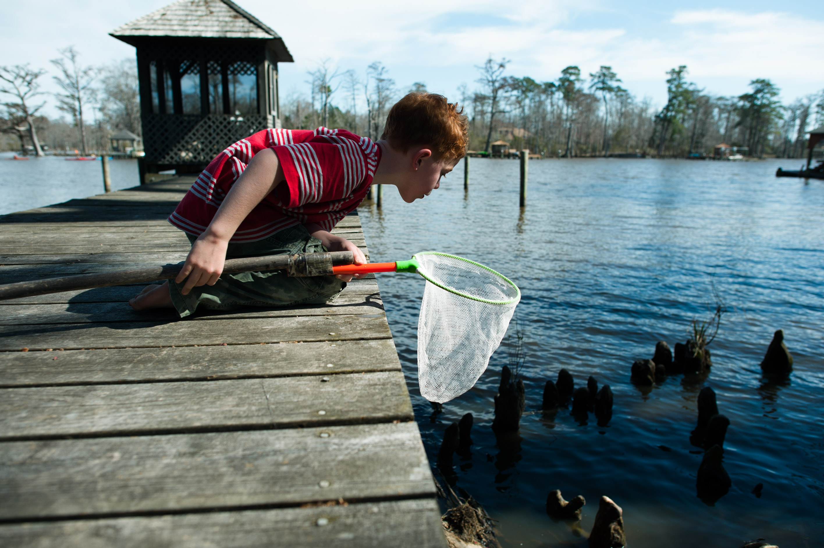 boy leans over dock with small fishing net - Documentary Family Photography