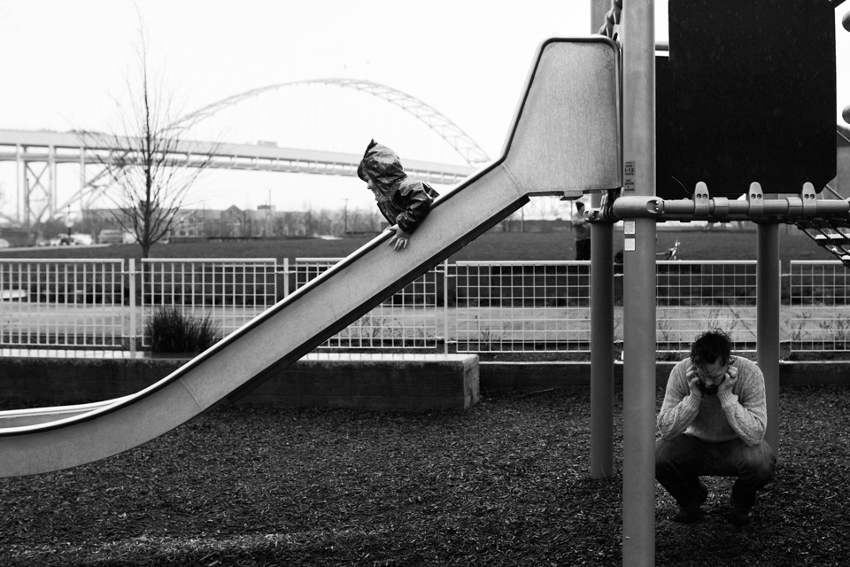 boy going down slide while dad talks on phone, documentary family photography