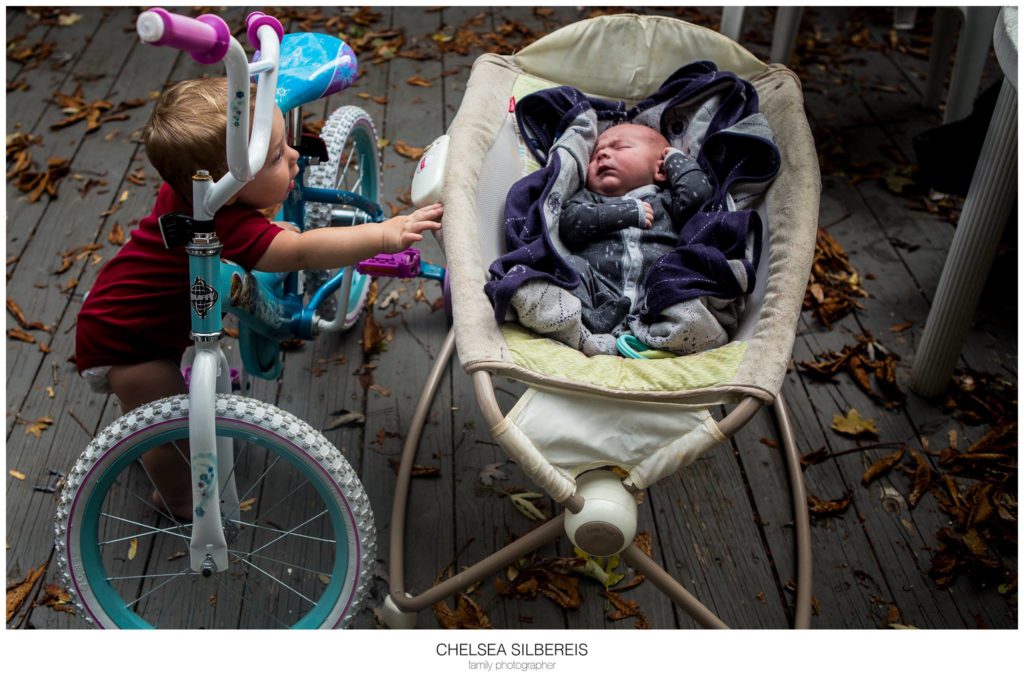child with bike looks on baby in rocker - Documentary Family Photography