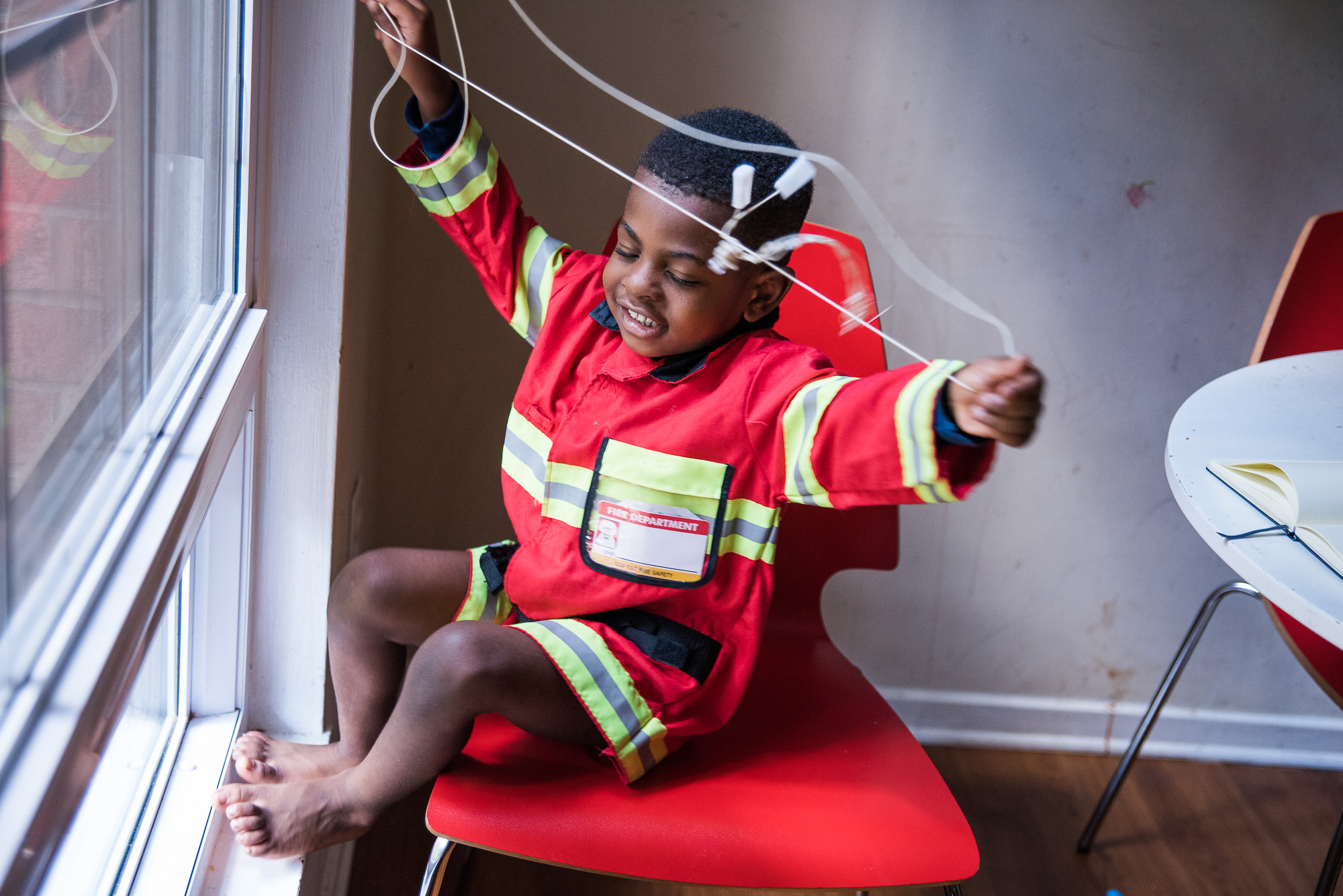 boy in fireman coat plays with mini blinds - Documentary Family Photography