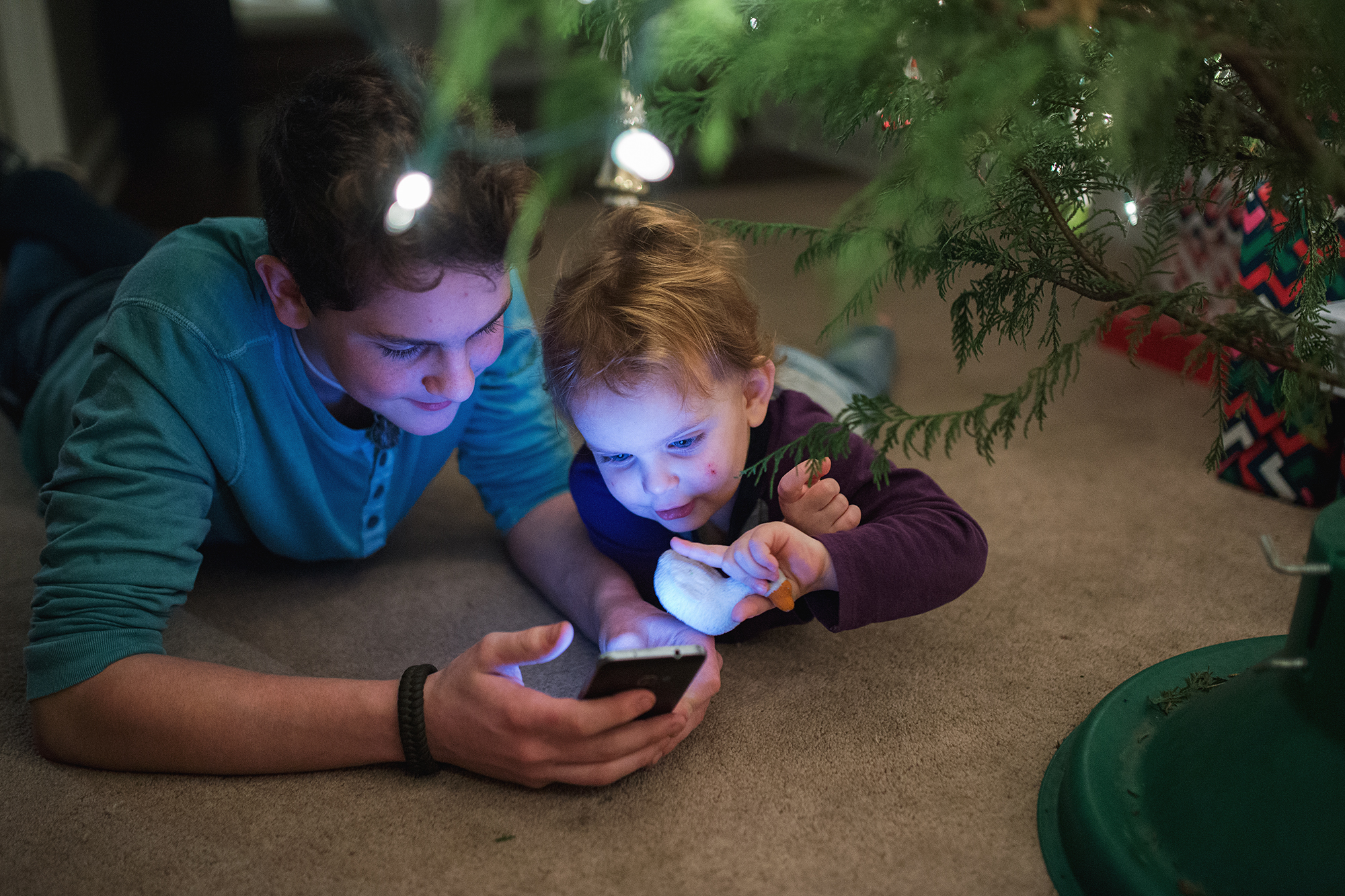 kids looking at phone under Christmas tree - Documentary Family Photography